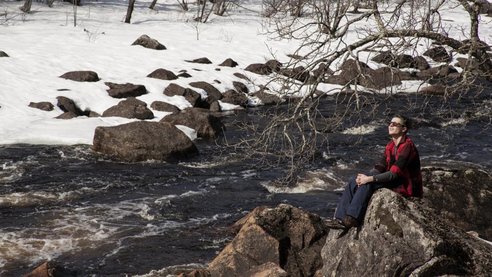 A person sits on a rock near rapids on a river.