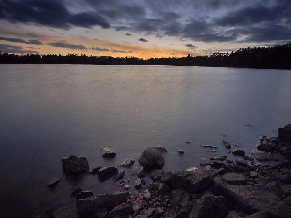 An image of a Boundary Waters lake right before sunrise, as the sun just begins to break through the clouds on the horizon