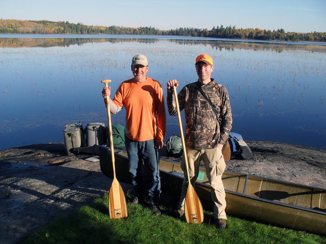Stu Osthoff and David Lien (right) stand with paddles near their canoe and gear by a wilderness lake.