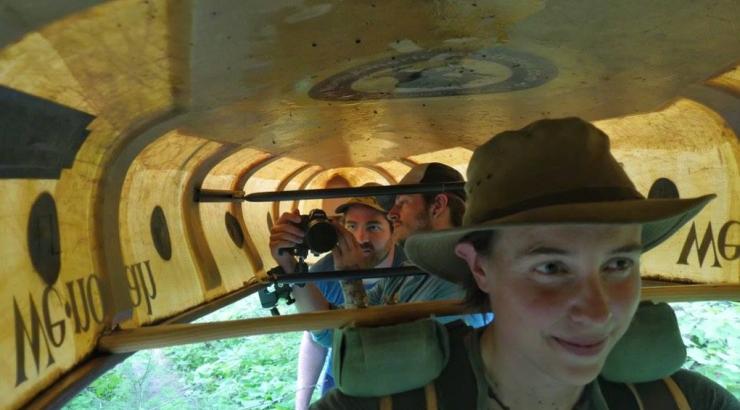 Amy Freeman portaging a canoe with 2 people under the canoe taking a photo of her