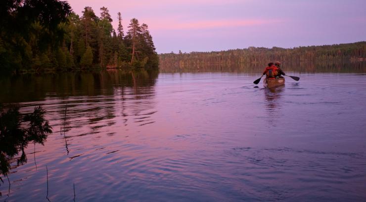 Dave and Amy Freeman sit in a canoe, paddling on a Boundary Waters lake at sunset as they look off-screen.