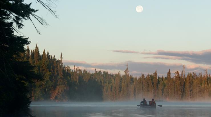 A canoe paddles away from the shore of a Boundary Waters lake during early morning, as a full moon hovers over them in a clear sky.