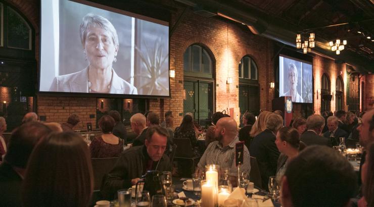 Photo of people eating dinner with candle in the center, with video of Sally Jewel being projected behind them