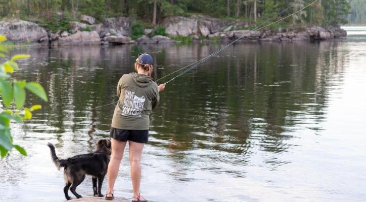 A person wearing a green "Save the Boundary Waters" windbreaker stands on a rock with their dog, who is a brownish color, as they cast a fishing rod into a Boundary Waters lake in the evening.