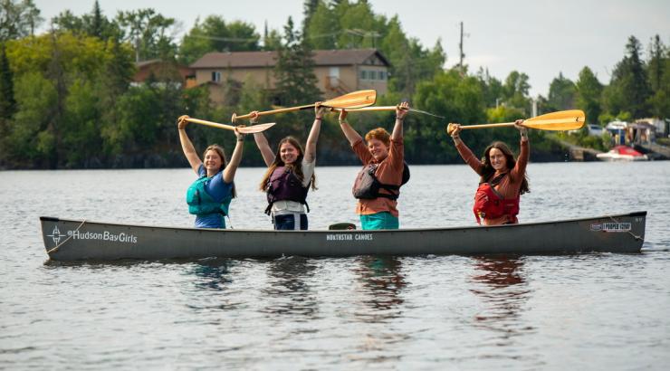 Four members of the Hudson Bay Girls sit in canoes with their paddles raised.