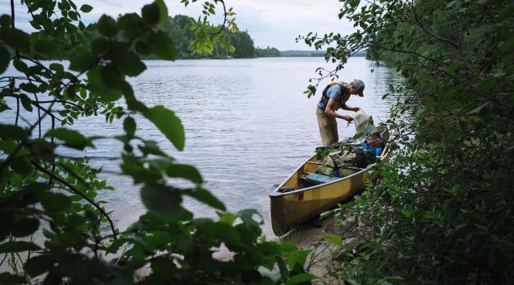 A man leans over his beige canoe on the shoreline of a Boundary Waters lake