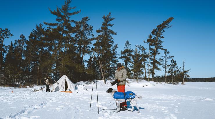 A man and dog play in the snow outside a canvas winter camping tent.