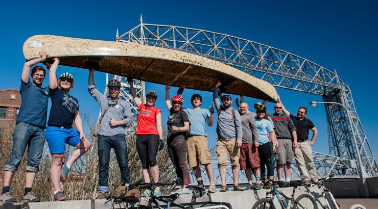 photo of 11 people holding up a tan Wenonah canoe infront of the Duluth Canal Park Bridge