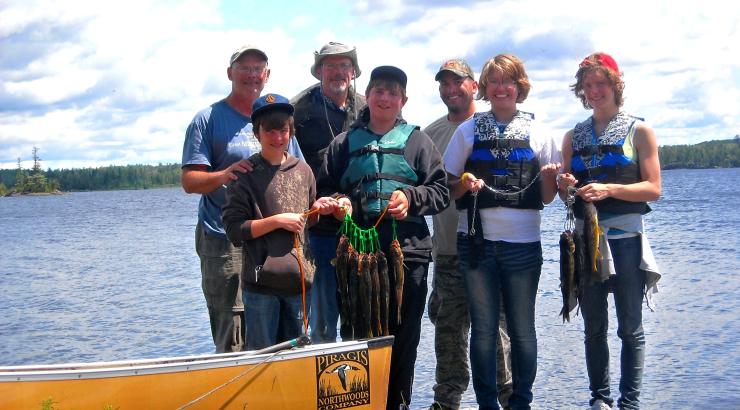 Photo of 7 people standing next to a canoe on the edge of a lake holding fish and smiling