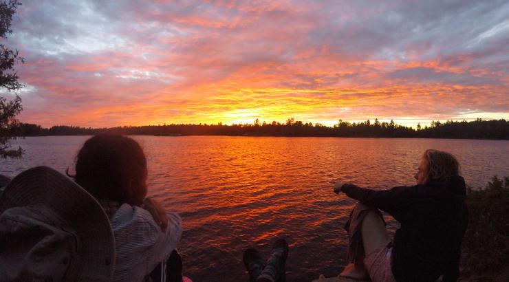 photo of 2 people sitting infront of a bright orange/pink sunset over water