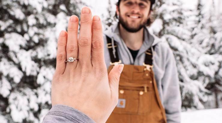 Photo of woman holding left hand out infront of camera with engagement ring on it with man standing behind hand smiling