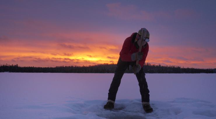 Person digging an ice hole on a lake infront of a Boundary Waters sunset