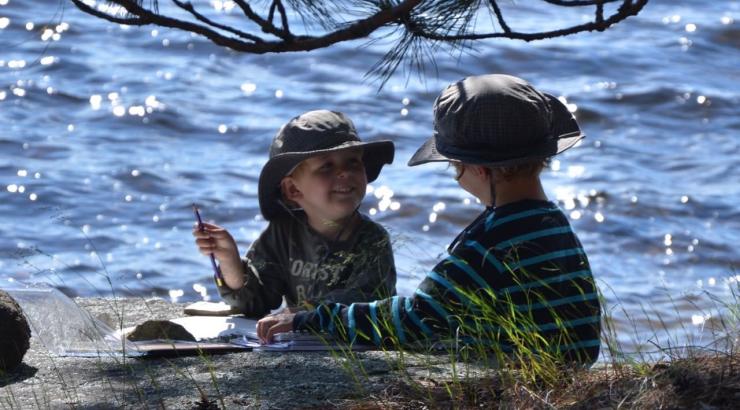 Photo of two children sitting by water in bucket hats under a tree smiling at eachother