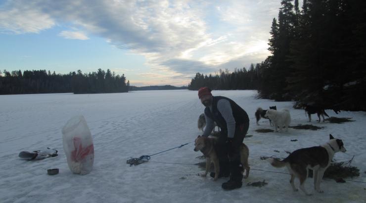 Photo of man standing on snow leaning over a bag with a dog next to him