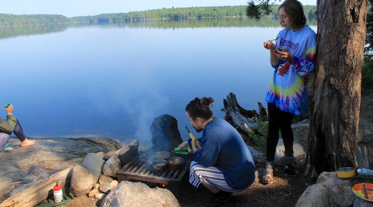 Photo of someone squatting nexto a campfire with someone next to them leaning against a tree with a lake in the background