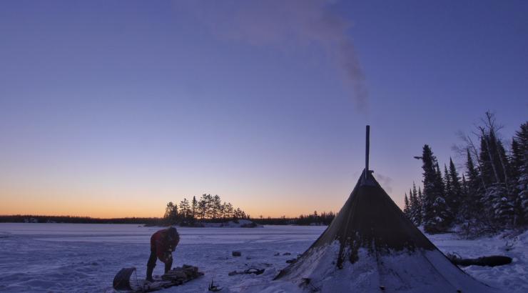 Photo of canvas tent silhouette and person leaning over sled on frozen lake infront of sunset