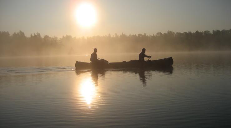 Silhouette of 2 people paddling canoe with sun shining in background