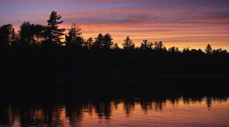 Photo of purple and orange sunset with tree silhouettes reflecting on the water