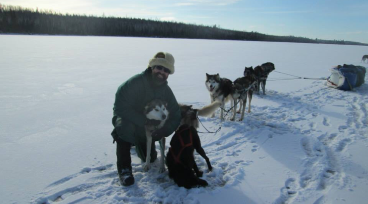 Photo of man kneeling on snow next to sled dogs