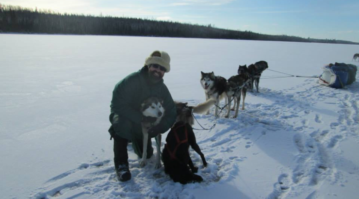 Photo of person kneeling next to a sled dog on a frozen lake with sled dogs in the background