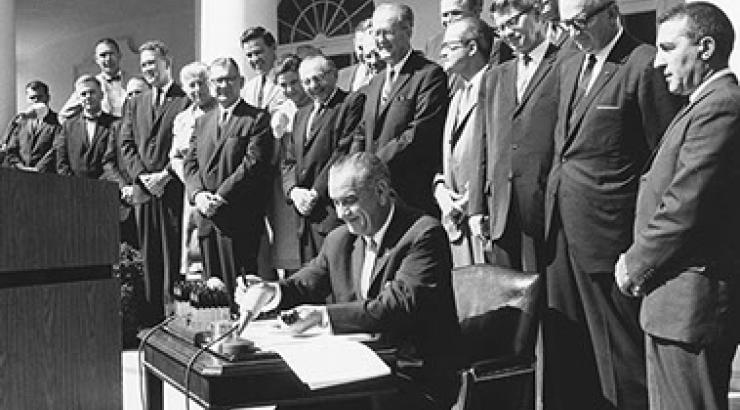 Black and white photo of Lynden B Johnson signing the Wilderness Act of 1964 with a row of men standing behind him in suits 
