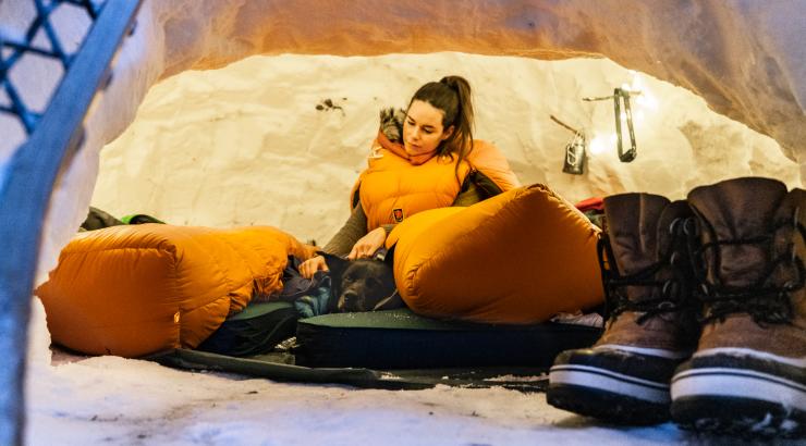 Photo of person sitting in an orange sleeping bag leaning against an igloo from the inside with dog laying next to them and big winter boots in the foreground