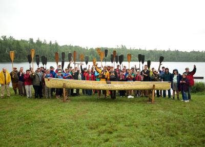 Community gathering holding up paddles behind a canoe. 