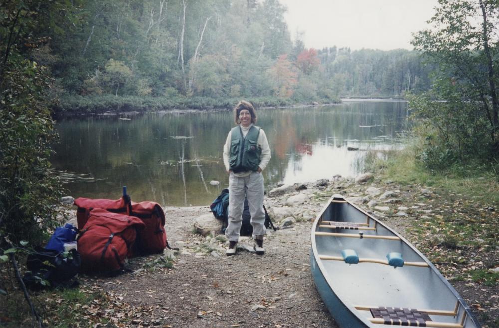 A woman poses between a portage pack and a canoe next to a Boundary Waters lake