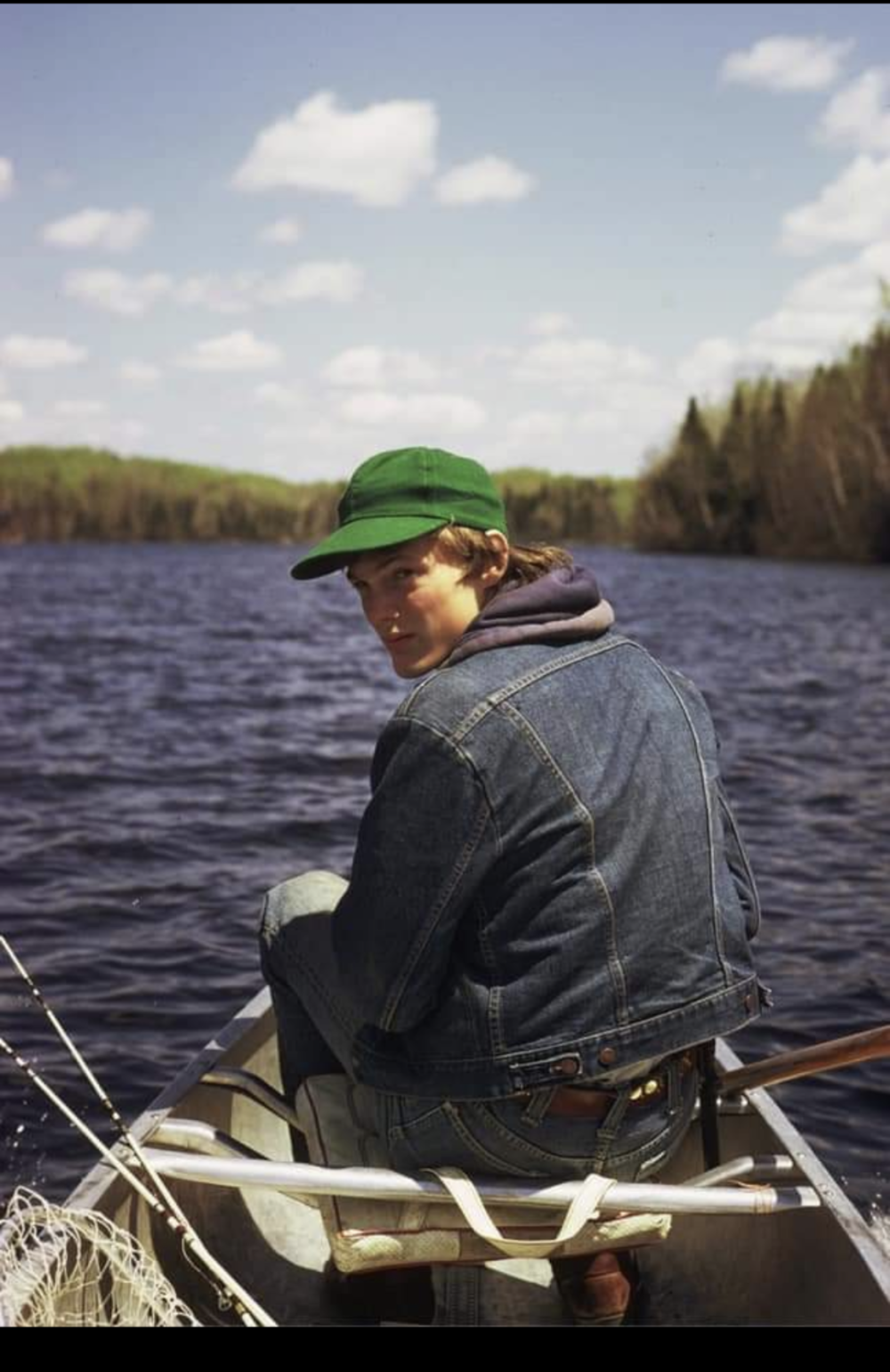 A man wearing a blue jean jacket and a green ball cap sits in the bow of a boat with his fishing equipment