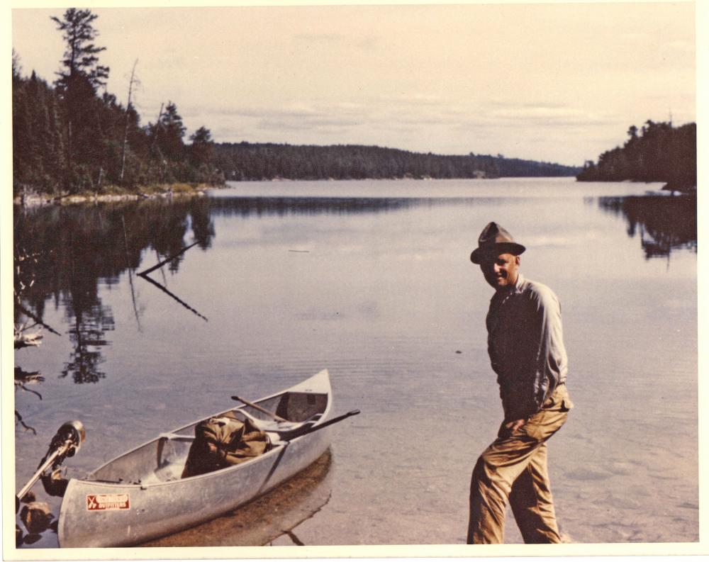 A man in a blue button up shirt and a grey sunhat poses next to a fully packed canoe in the Boundary Waters