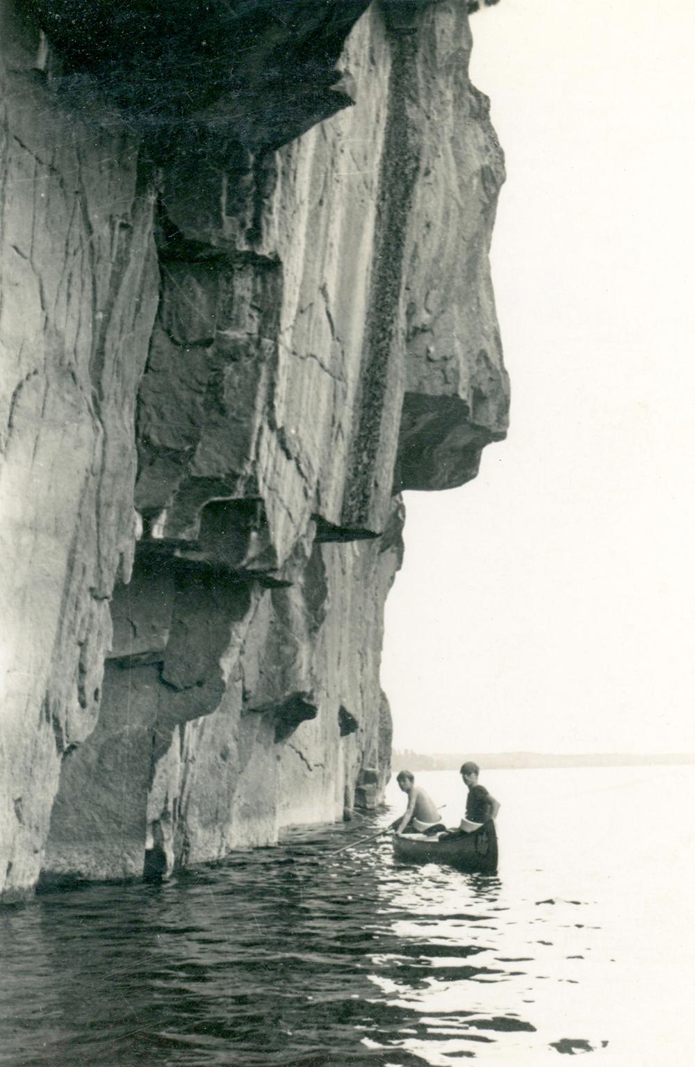 Two young men paddle in a canoe directly next to a cliff, which rises out of a Boundary Waters lake