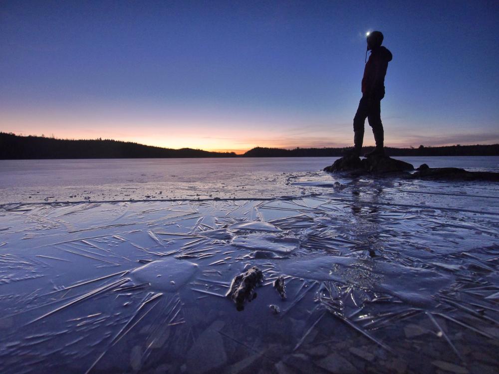 A man stands on a rock that juts out onto a frozen lake