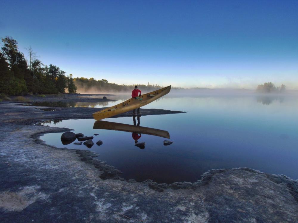 A person on a shore lifts their canoe out of a lake in the Boundary Waters