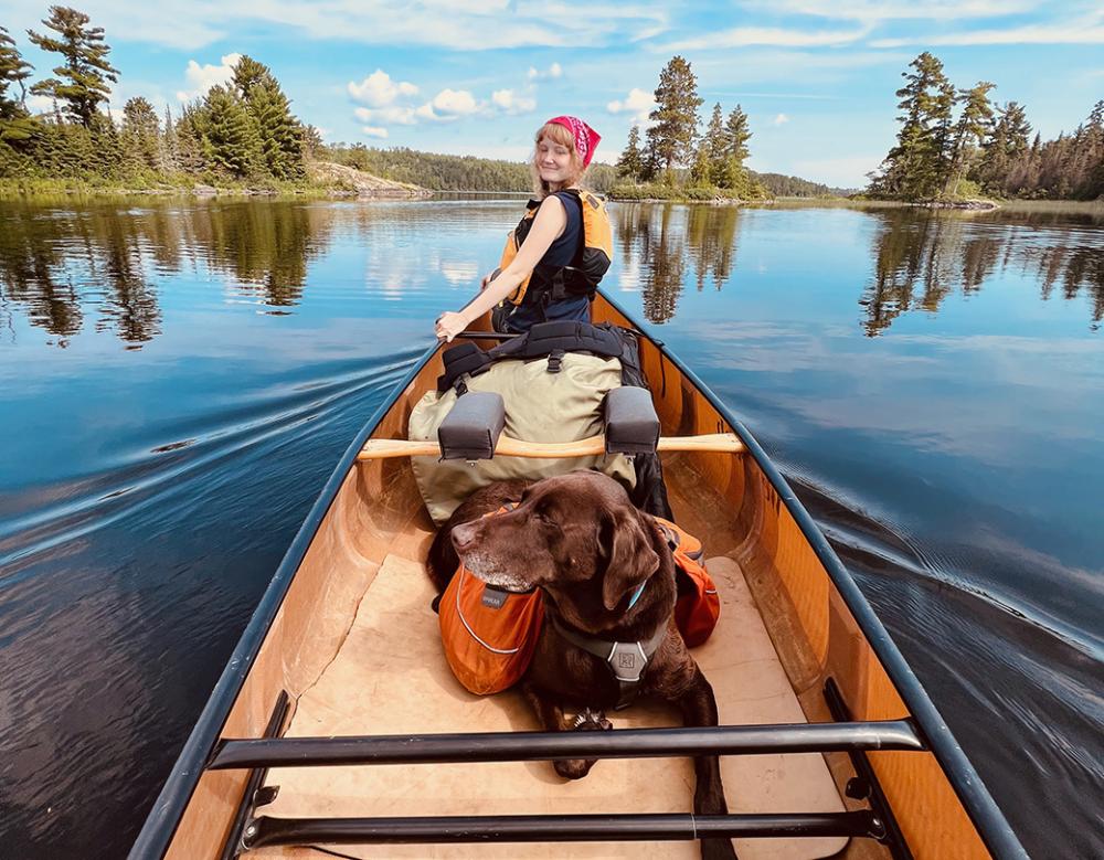 A brown dog rests in the bottom of the canoe on a lake. 