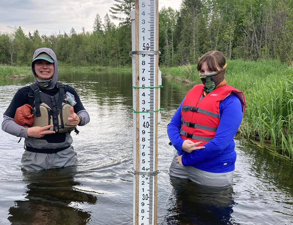 Two researchers stand in a creek next to a water gauge. 