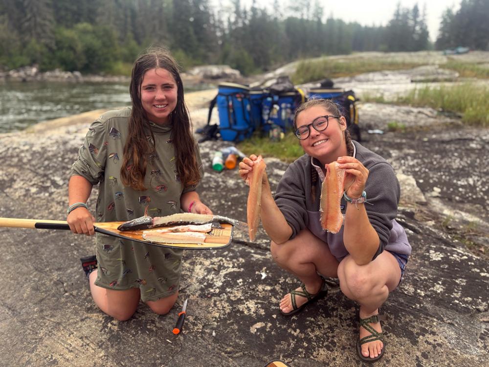 Two crew members of the Hudson Bay Girls fillet a fish near a waterfall.