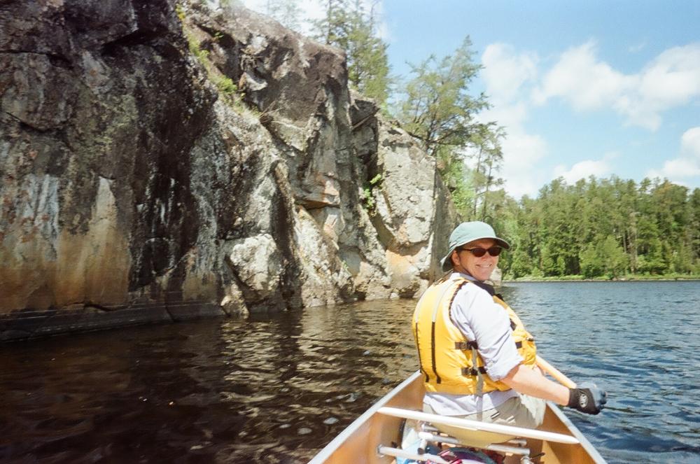 A woman turns around in the bow of a canoe to pose while paddling past a small cliff jutting out of a lake in the Boundary Waters