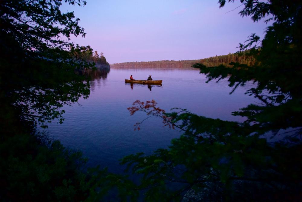 Through an opening in the trees, two people are photographed canoeing on a Boundary Waters lake right before sunrise