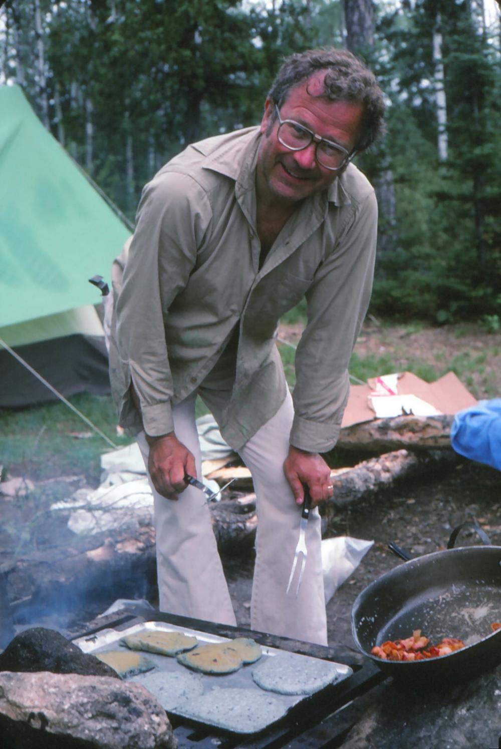 A man in a tan button-up shirt poses as he works on flipping blueberry pancakes over the fire