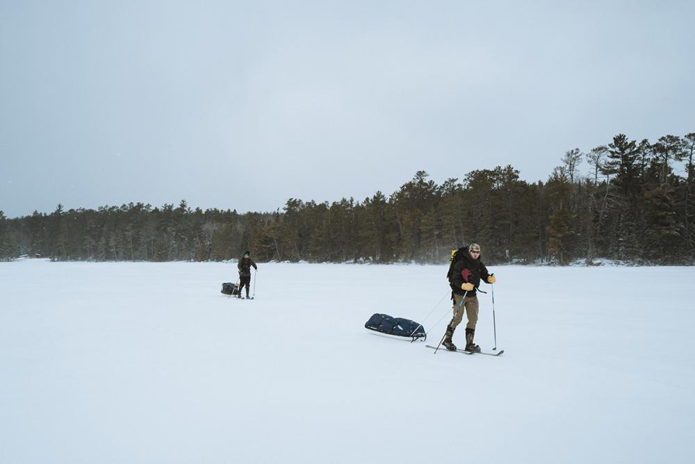 Two people pull winter sleds across lake ice in the wilderness.