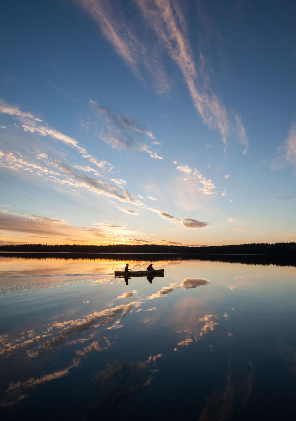 Two people canoe on a glassy Boundary Waters lake, which reflects the clouds and sunset on the horizon