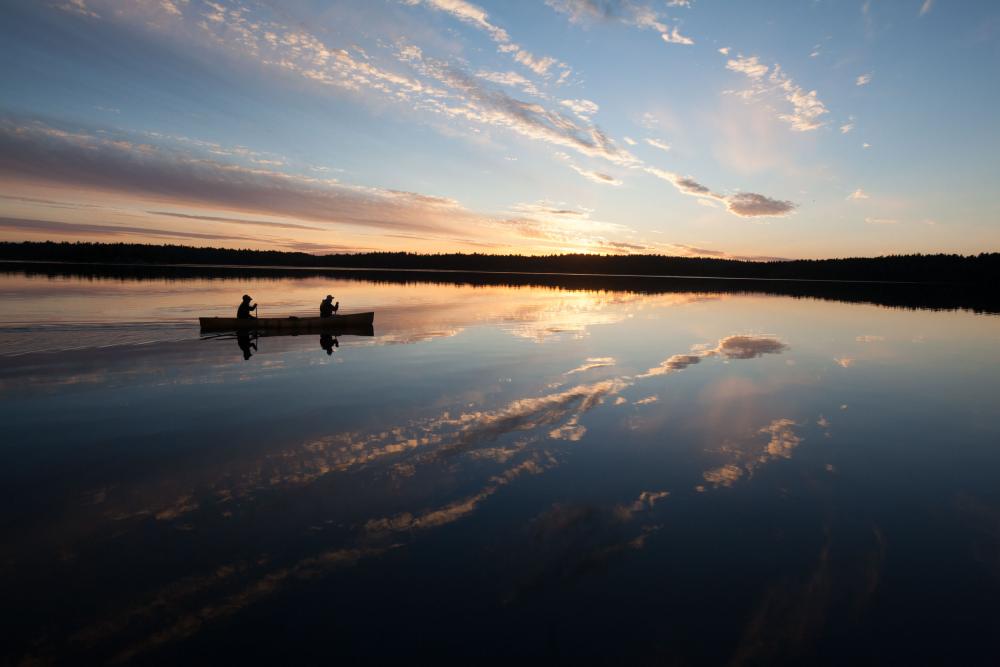 Two people canoe on a glassy Boundary Waters lake, which reflects the clouds and sunset on the horizon