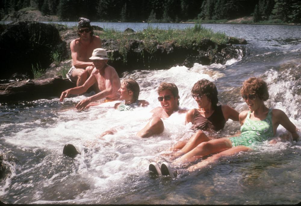 On a summer day, 6 people sit in a shallow Boundary Waters section of rapids to cool off