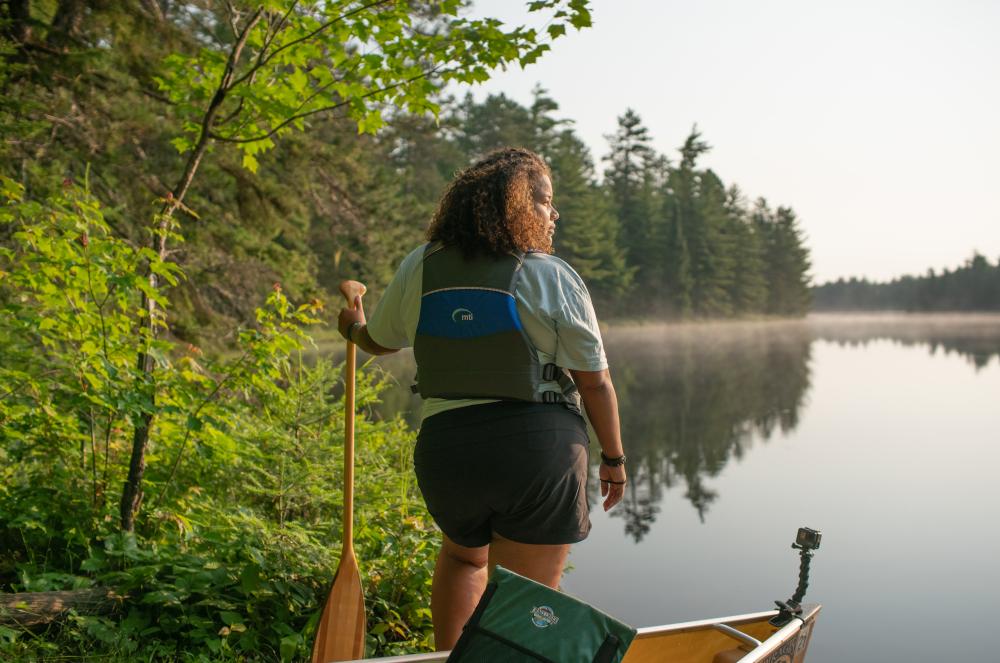 A person with a canoe paddle and lifejacket stands near a canoe on a lake shore, ready to set out for a day of paddling
