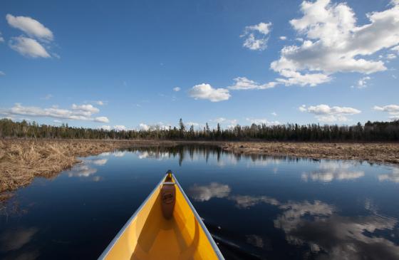 Photo of front of canoe on water with white clouds reflecting over Boundary Waters