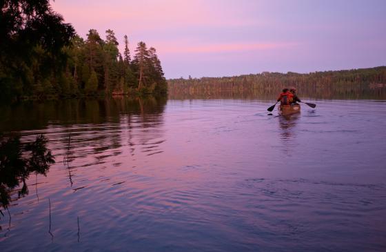 Dave and Amy Freeman sit in a canoe, paddling on a Boundary Waters lake at sunset as they look off-screen.