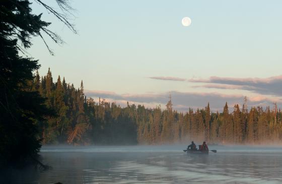 A canoe paddles away from the shore of a Boundary Waters lake during early morning, as a full moon hovers over them in a clear sky.