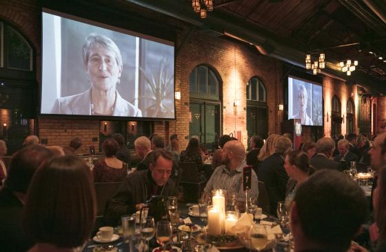 Photo of people eating dinner with candle in the center, with video of Sally Jewel being projected behind them