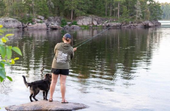 A person wearing a green "Save the Boundary Waters" windbreaker stands on a rock with their dog, who is a brownish color, as they cast a fishing rod into a Boundary Waters lake in the evening.