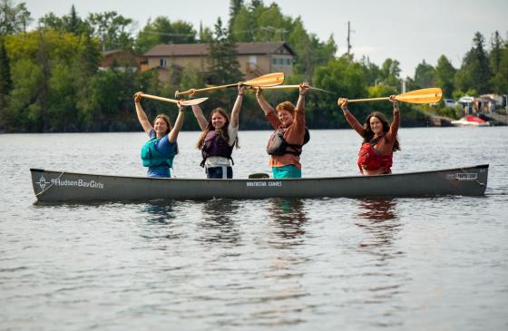 Four members of the Hudson Bay Girls sit in canoes with their paddles raised.
